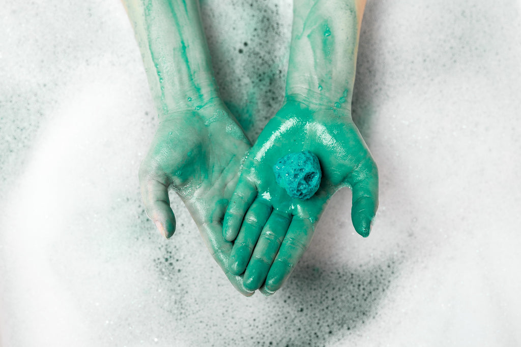 Blue and yellow bath bomb on person's hand melting in water and turning the water to blue