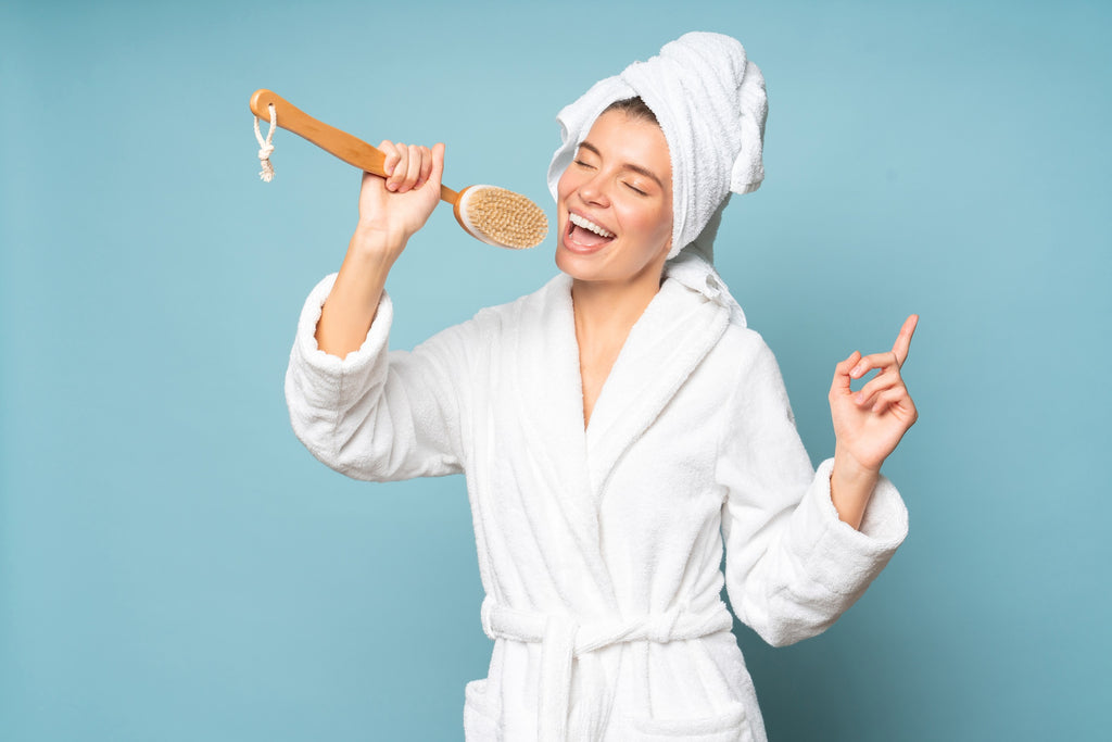 Woman wearing bathrobe and wrapped towel on head using a long-handed body scrub brush as a mic to sing